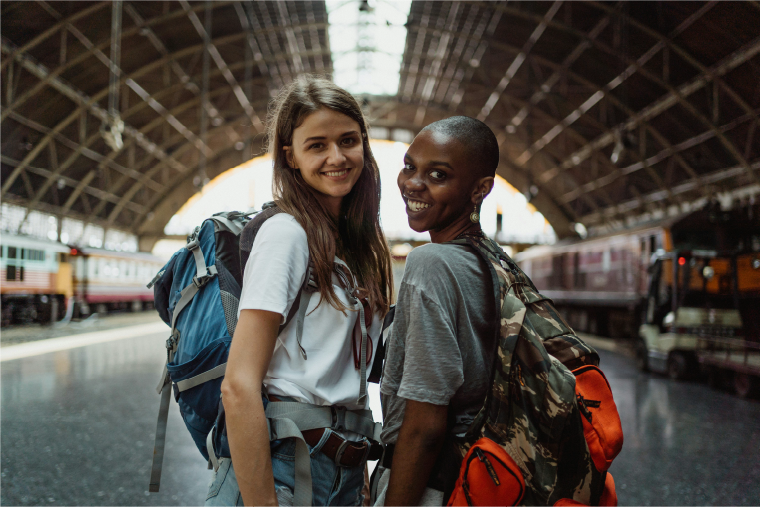 Dos mujeres con mochilas en una estación de tren, listas para una nueva aventura.